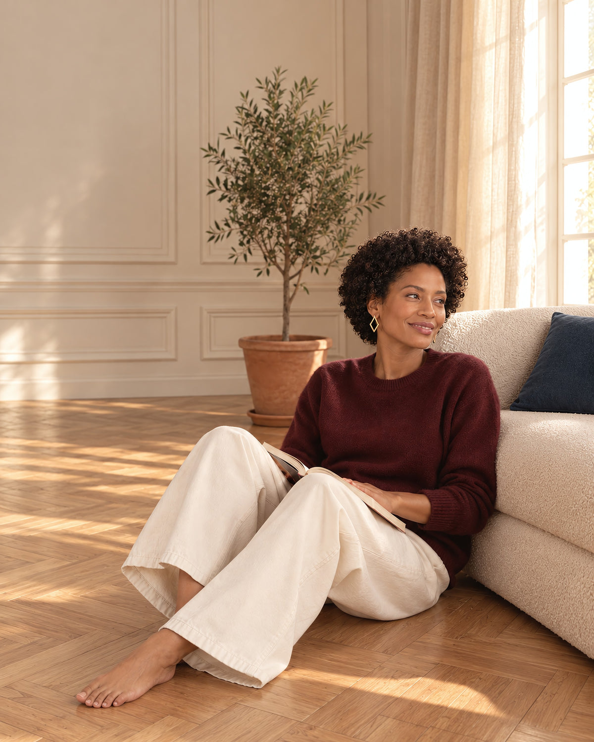 Femme en pull bordeaux et pantalon écru, assise sur le parquet contre le canapé, un livre ouvert