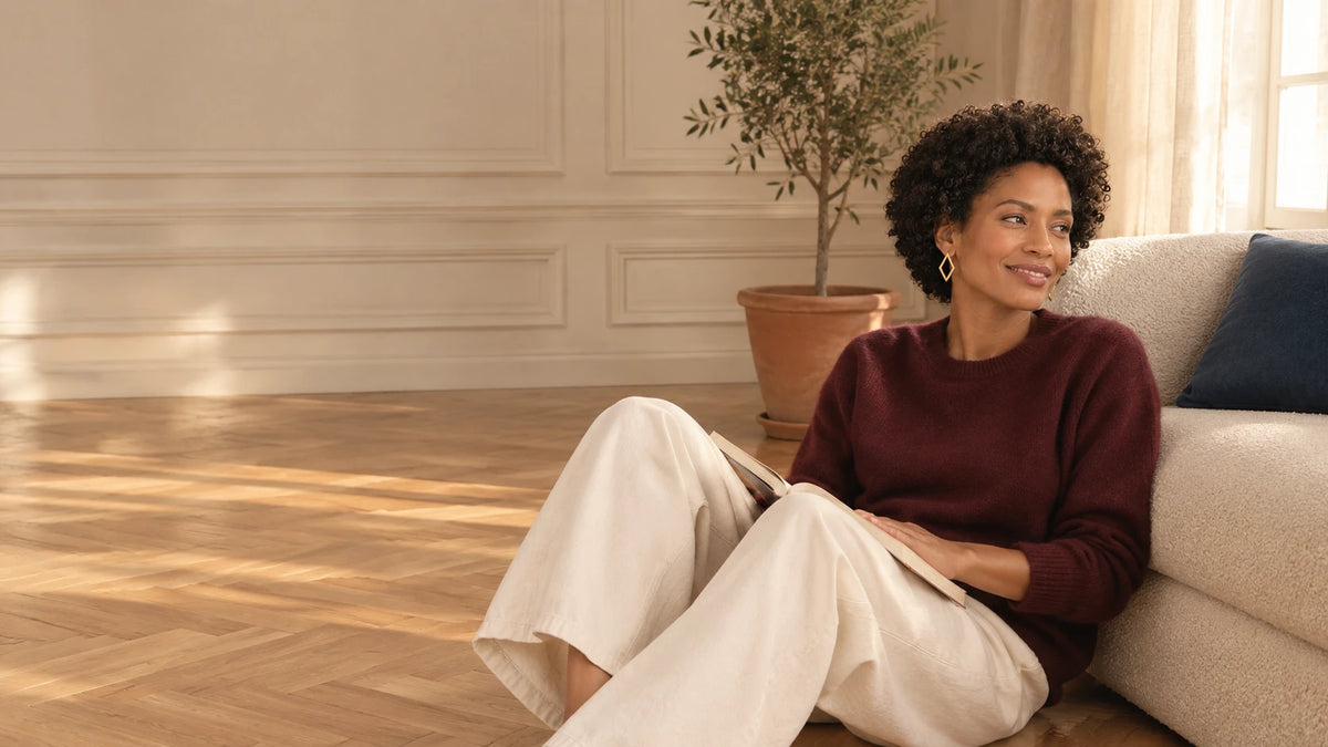 Femme en pull bordeaux et pantalon écru, assise sur le parquet contre le canapé, un livre ouvert